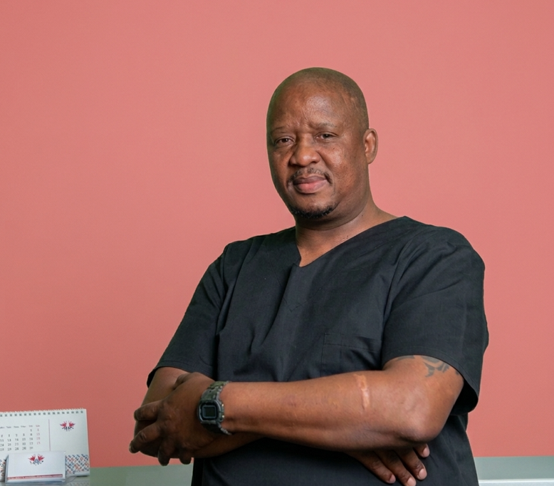 Macdonald Molaolwa, the doctor's secretary at Mahikeng Medical Center, smiling with arms crossed in black scrubs